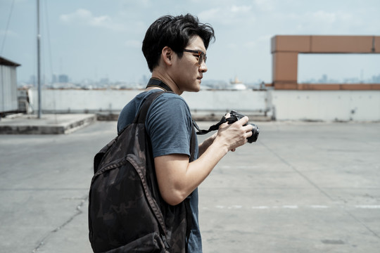 年轻英俊的亚洲摄影师戴着太阳镜手持相机站在屋顶上。