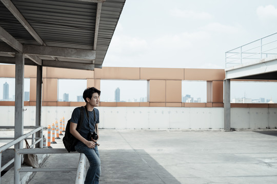 年轻英俊的亚洲摄影师坐在屋顶上。城市街头摄影。