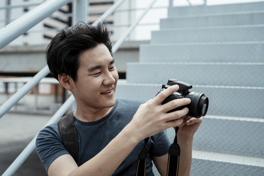年轻英俊的亚洲摄影师坐在楼梯上拿着相机在屋顶拍照。