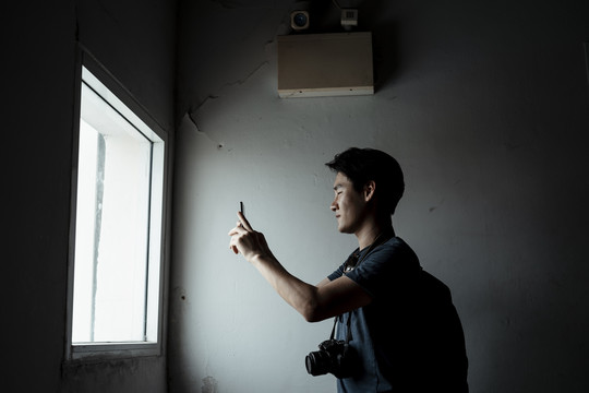 年轻英俊的亚洲摄影师男子在大楼内用智能手机在窗口拍照。