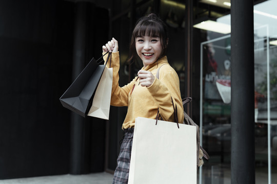 兴高采烈的亚洲年轻女子穿着黄色毛衣，拿着许多纸袋在购物中心外散步。