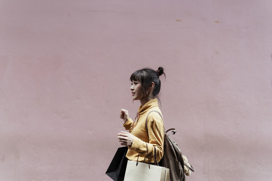 侧视图：穿着黄色毛衣的亚洲妇女手拿纸袋孤立地站在粉色背景上-复印空间。