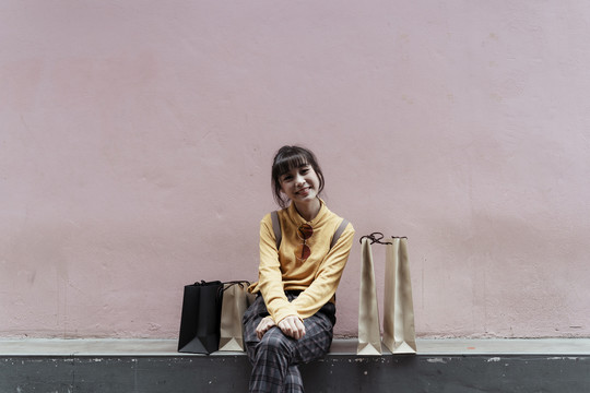 快乐的亚洲可爱女人坐在地板上，手拿粉色背景的购物纸袋。