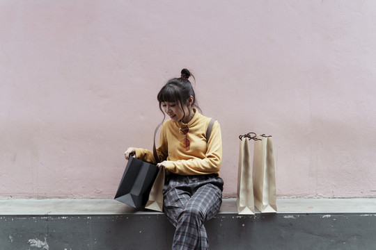 快乐的亚洲女人穿着黄色毛衣坐在地板上，在粉色背景的黑色购物袋里寻找东西。