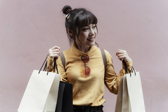 一位穿着黄色毛衣，背着粉色背景的购物袋的快乐的亚洲年轻女子。