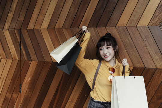 穿着黄色衬衫的兴奋美丽的亚洲妇女的肖像，在木制背景上拿着购物纸袋。