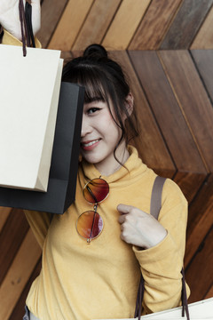 身着黄色毛衣、手持购物纸袋、站在木制背景上的快乐亚洲妇女的肖像。