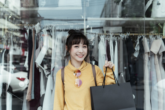 一位穿着黄色毛衣、手持黑色购物袋的亚洲美女站在服装店门口。