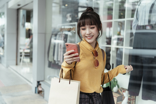 穿着黄色毛衣的泰国亚裔美女在购物店前的大街上走着，用智能手机享受着网上购物的乐趣。