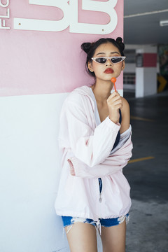 年轻迷人的亚泰女性深色双髻发型，粉色头巾，戴着时尚眼镜，手里拿着棒棒糖在停车场。