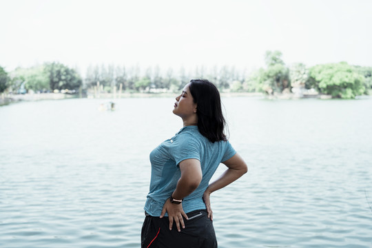 侧视图-胖乎乎的亚洲女人在湖边伸展身体，在大自然中慢跑前热身。