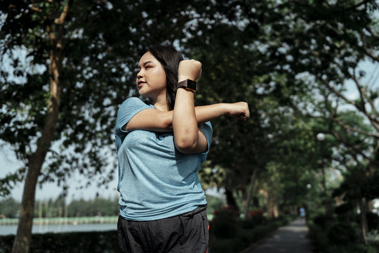 胖乎乎的运动型亚洲女子，穿着蓝色运动服，在公园跑步前伸展手臂。