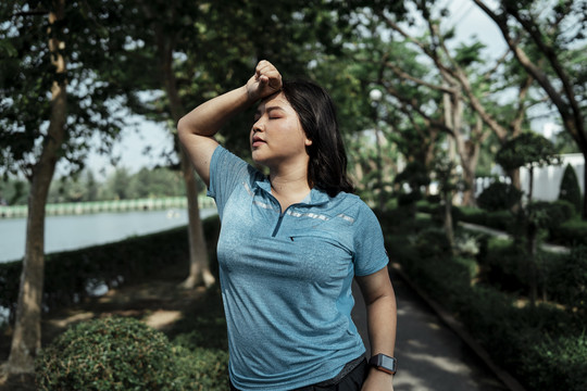 穿着蓝色运动服的疲惫胖胖的亚洲女人在公园跑步后休息。胖女孩在公园锻炼。
