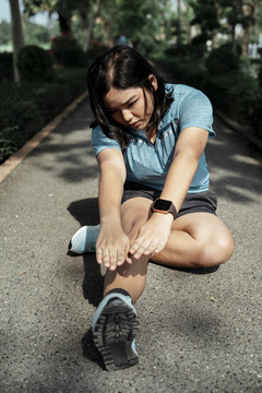 胖乎乎的运动型亚洲女子，穿着蓝色运动服，在公园的跑道上伸展双腿。
