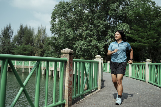 胖乎乎的运动型亚洲女人穿着蓝色运动服在湖边的自然公园跑道上跑步。