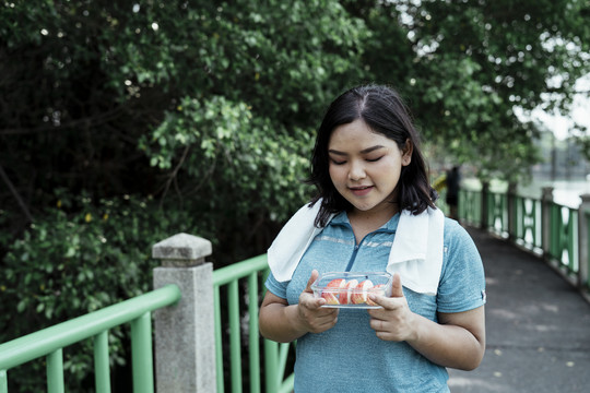 一个胖乎乎的运动型亚洲女人，穿着蓝色运动服，手里拿着一个装满苹果片的玻璃碗。