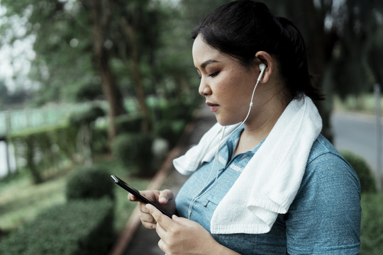 胖乎乎的运动型亚洲女人穿着蓝色运动服，用智能手机的耳机听音乐。