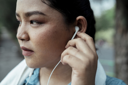 特写胖乎乎的运动型亚洲女人穿着蓝色运动服，一边在公园里锻炼一边用耳机听音乐。