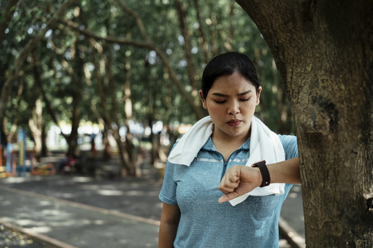 胖乎乎的运动型亚洲女人穿着蓝色运动服，看着公园里的智能手表。