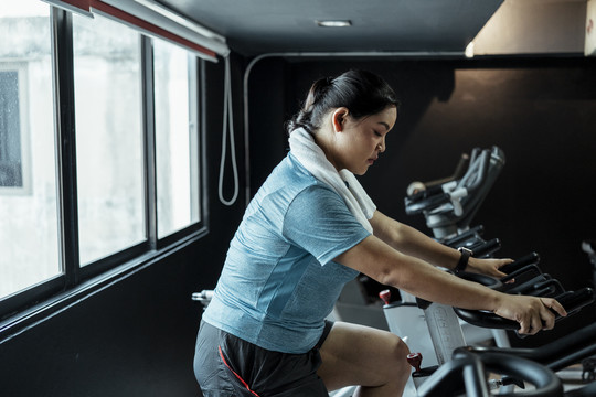胖乎乎的运动型亚洲女子，穿着蓝色运动服，在健身房的电动自行车上锻炼。