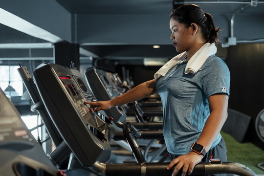 胖乎乎的运动型亚洲女子，穿着蓝色运动服，在体育馆内的跑步机上跑步。