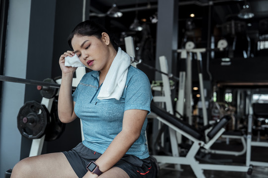 穿着蓝色运动服的疲惫胖乎乎的运动型亚洲女人在健身房辛苦锻炼后，在长凳上放松。