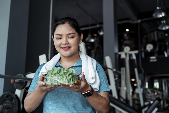 胖乎乎的运动型亚洲女人穿着蓝色运动服在健身房吃蔬菜健康午餐。