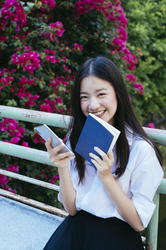 漂亮的深色长发年轻大学生身着校服手持一本书和一部智能手机。