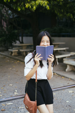 美丽的深色长发年轻大学生身着校服在公园里拿着一本书。