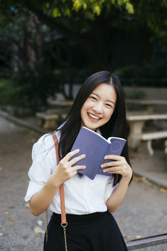 美丽的深色长发年轻大学生身着校服在公园里拿着一本书。
