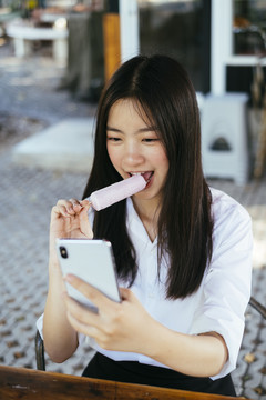漂亮的深色长发年轻大学生穿着校服用智能手机和吃冰激凌棒。