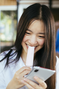 漂亮的深色长发年轻大学生穿着校服用智能手机和吃冰激凌棒。