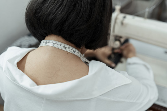 后视图-年轻的泰国设计师裁缝在缝纫机站。把卷尺挂在她脖子上。
