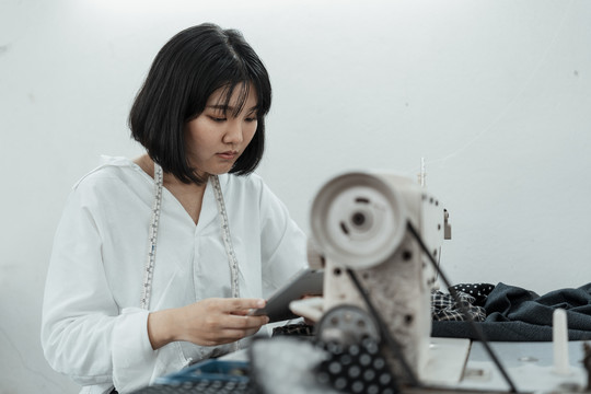 年轻的亚洲设计师坐在缝纫站的桌子旁，用智能手机在网上寻找灵感。
