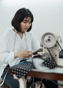 年轻的亚洲设计师坐在缝纫站的桌子旁，用智能手机在网上寻找灵感。