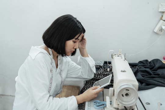 年轻的亚洲设计师在白色的房间里，在缝纫机台前工作，看着平板电脑，头痛不已。
