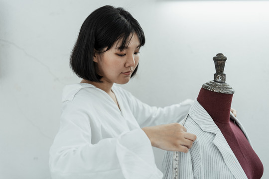 在白色展厅里，年轻的亚洲设计师正在用卷尺在假人肩膀上做裙子。