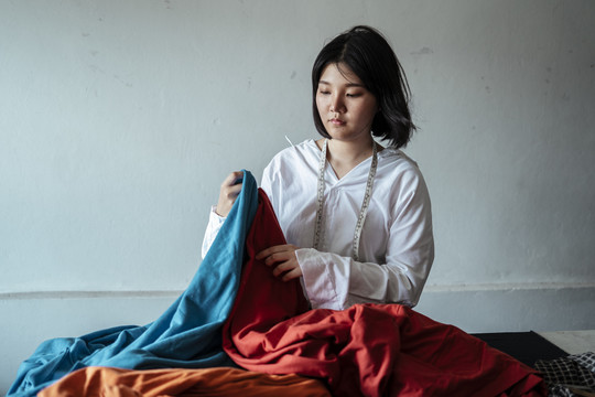 一位年轻的亚洲设计师，一头短发，穿着白衬衫，从一堆布料中挑选布料来做衣服。