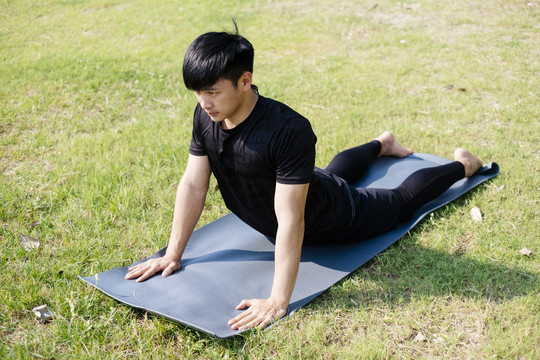 一位身穿黑色运动服的亚泰男子在绿地的垫子上做瑜伽。户外。