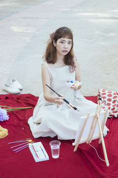 美丽的亚泰艺术家穿着白色的衣服，在红色的垫子上用画笔在画架上作画。
