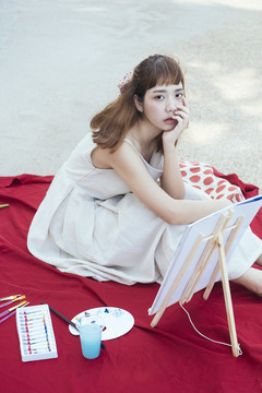 美丽的亚泰艺术家穿着白色的裙子坐在红色的垫子上，用画架和绘画的东西。