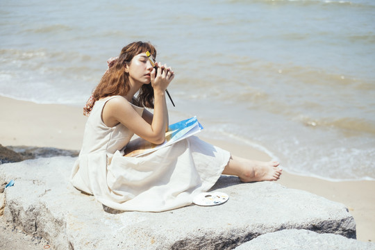 夏天，美丽的亚泰艺术家穿着白色连衣裙，坐在海滩的岩石上，用帆布做装饰。