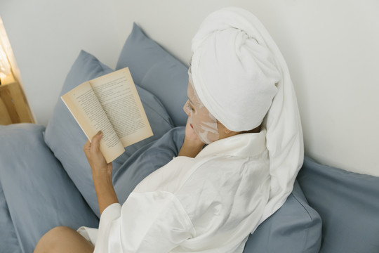 后视图-穿着白色浴袍的年轻女子戴着化妆面具躺在卧室里看书。