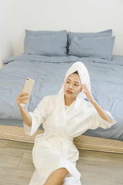 穿着白色浴袍的年轻女子在卧室里用智能手机自拍。