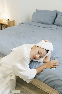 穿着白色浴袍的年轻女子戴着化妆面具睡在床上，卧室里拿着智能手机。