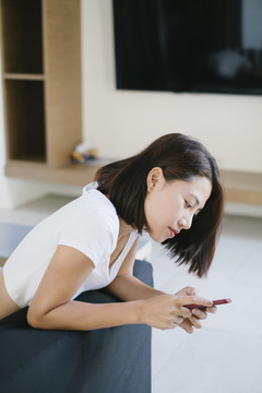 性感的亚泰女人穿着白衬衫躺在客厅的沙发上用智能手机。