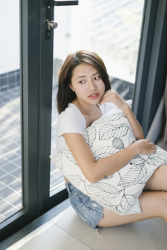 性感的亚泰女人穿着白衬衫抱着枕头坐在窗前。