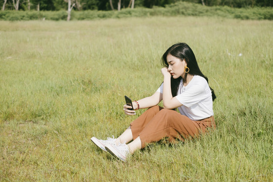 穿着休闲服的黑色长发女子坐在草地上，用智能手机自拍。