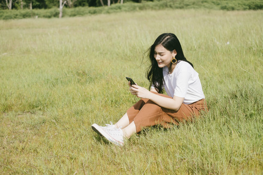 穿着休闲服的黑色长发女子坐在草地上，用智能手机自拍。