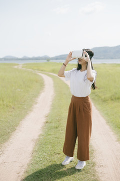 黑色长发女子穿着休闲服，戴着VR护目镜，在泥泞的草原上行走，享受虚拟现实技术。